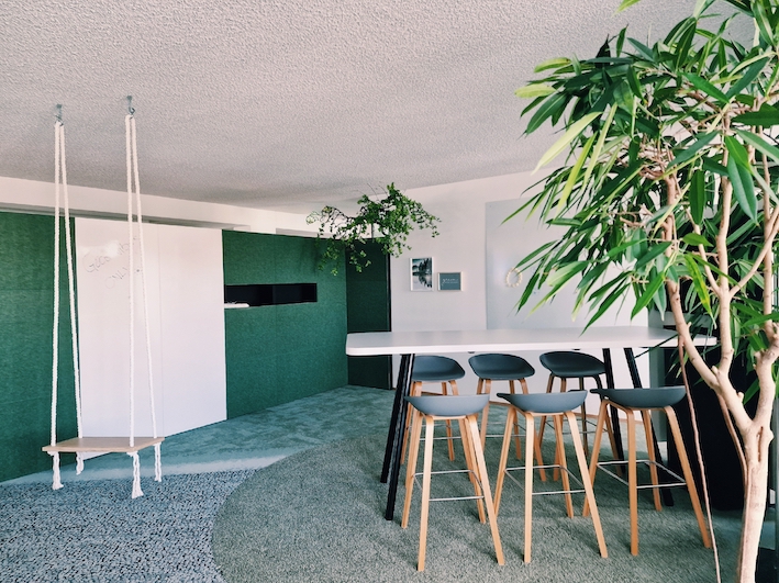 Ein grüner Kreativraum mit Schaukel, Interface Teppich, einem Hochtisch, Screen, Whiteboards und Hochstühlen. Perfekt für inspirierende Meetings und innovative Besprechungen.