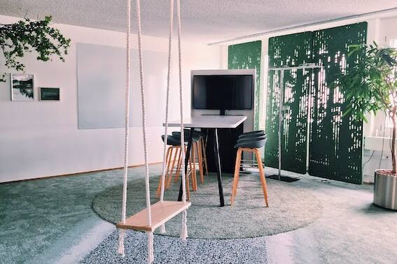 Ein grüner Kreativraum mit Schaukel, Interface Teppich, einem Hochtisch, Screen, Whiteboards und Hochstühlen. Perfekt für inspirierende Meetings und innovative Besprechungen.